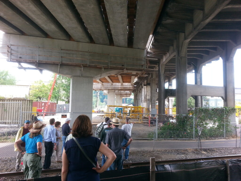 This picture, looking west from the east bank of the river, is a good look at the construction tricks used to keep traffic moving during the bridge construction.  The lighter concrete on the left is the new construction, while the darker parts to the right are the old bridge.  This part of the old bridge is carrying traffic right now.  You’ll notice that the closest arch on the new side is a “T”, but look further down the bridge… you can barely see it, but the farther new arches have two supporting columns instead of just one.  When the rest of the bridge is complete, this will be one of the last areas to be addressed.  Traffic will be completely diverted onto the new construction while this last remaining part of the old bridge is demolished, and the “T” arches of the new construction will be completed with their second supports.